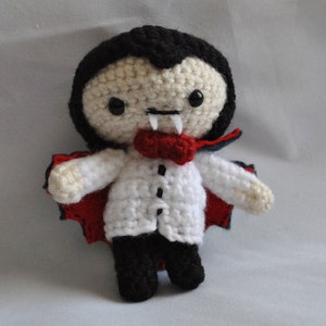 May include: A crocheted plush toy vampire with a red cape, black hair, and white shirt. The toy has a friendly expression with large eyes and a small mouth.