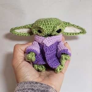 May include: A crocheted green and purple baby Yoda wearing a purple sweater. The toy is being held in a person's hand.