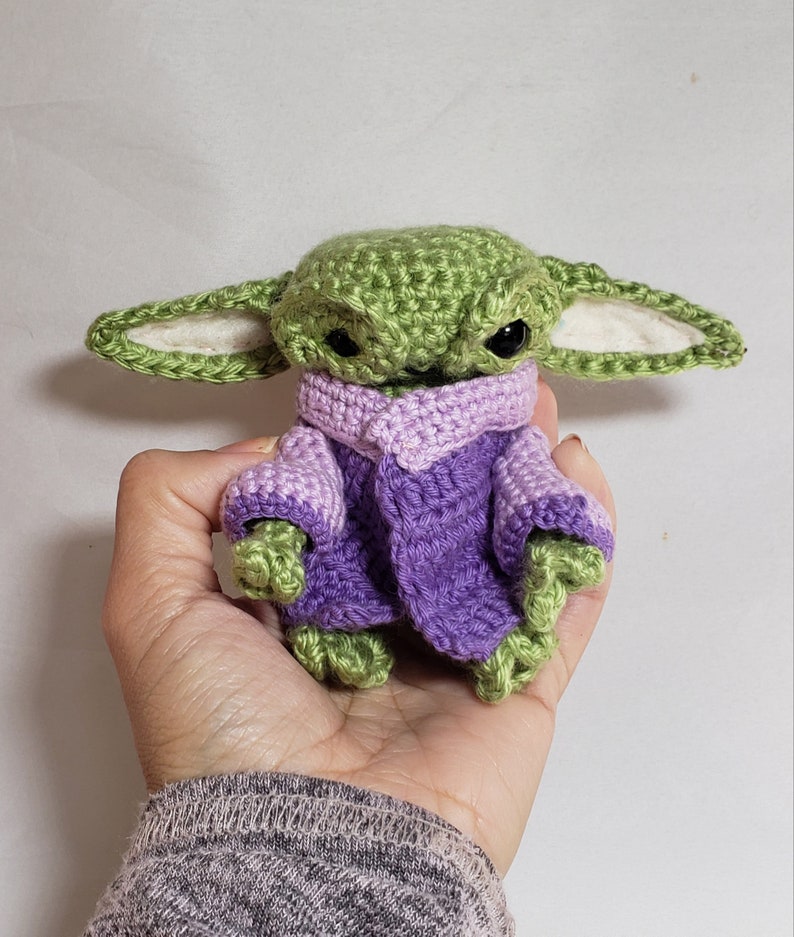 May include: A crocheted green and purple baby Yoda wearing a purple sweater. The toy is being held in a person's hand.