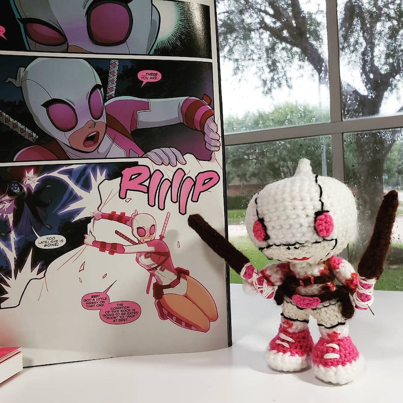 May include: A crocheted amigurumi doll of Deadpool in a pink and white color scheme. The doll is holding two brown swords and is standing in front of a comic book.
