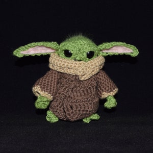 May include: A green crocheted plush toy with large ears, brown eyes, and a brown crocheted coat. The toy is standing on a black surface.
