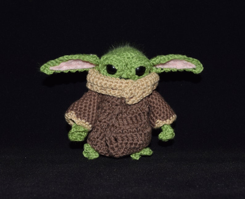 May include: A green crocheted plush toy with large ears, brown eyes, and a brown crocheted coat. The toy is standing on a black surface.