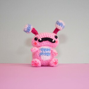 May include: A pink crocheted plush toy with blue and white striped accents. The toy has a black moustache and large black eyes.