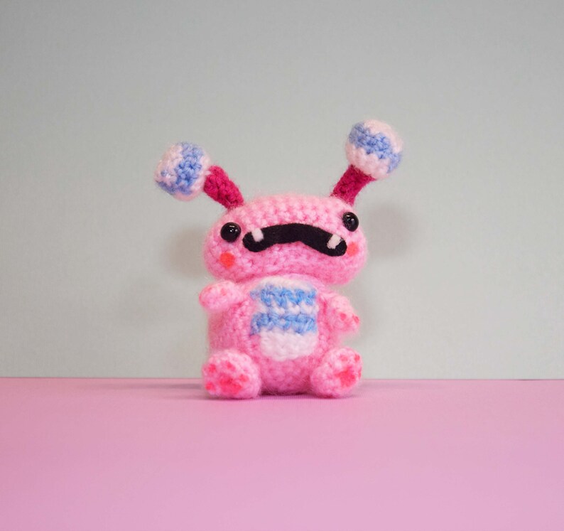 May include: A pink crocheted plush toy with blue and white striped accents. The toy has a black moustache and large black eyes.