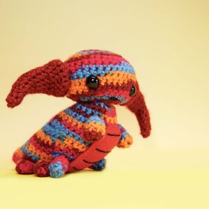 May include: A colorful crocheted stuffed animal with a long snout and large eyes. The animal is made with red, orange, blue, and yellow yarn.