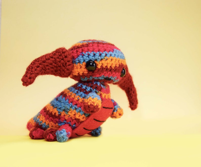 May include: A colorful crocheted stuffed animal with a long snout and large eyes. The animal is made with red, orange, blue, and yellow yarn.