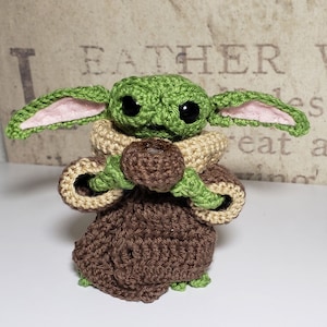 May include: A crocheted green and brown baby Yoda doll holding a brown chocolate chip cookie. The doll has large eyes and pink ears.