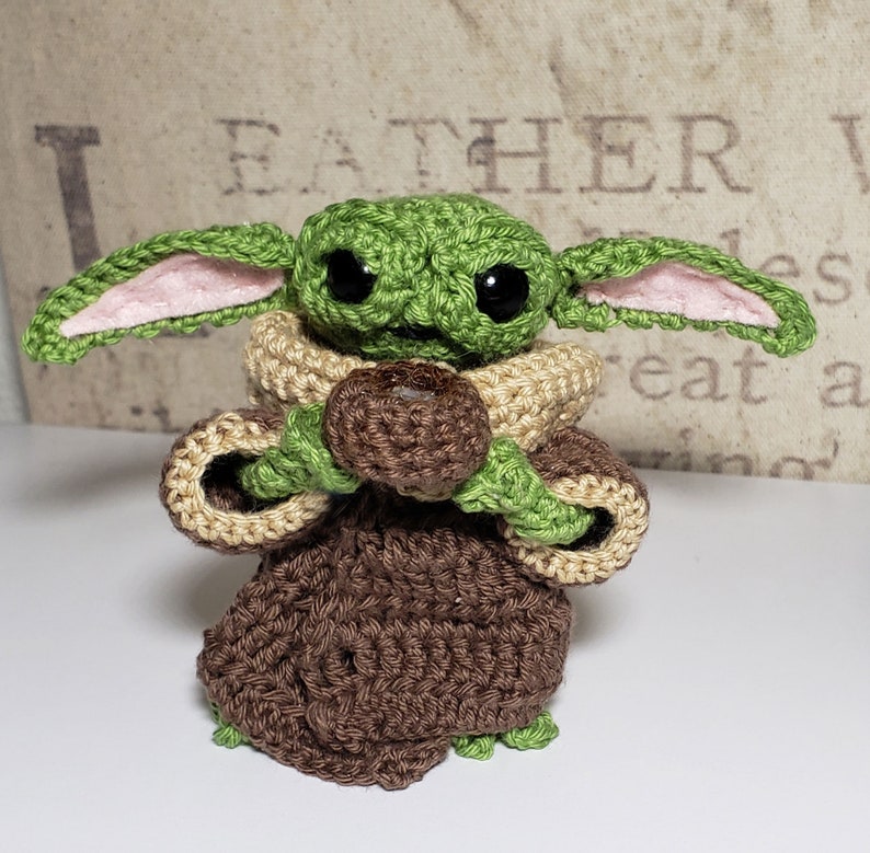 May include: A crocheted green and brown baby Yoda doll holding a brown chocolate chip cookie. The doll has large eyes and pink ears.