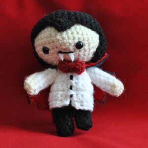 May include: A crocheted plush toy vampire with a red cape, white shirt, and black bow tie. The toy has black hair, a white face, and fangs.