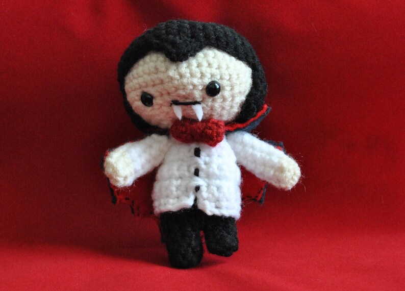 May include: A crocheted plush toy vampire with a red cape, white shirt, and black bow tie. The toy has black hair, a white face, and fangs.