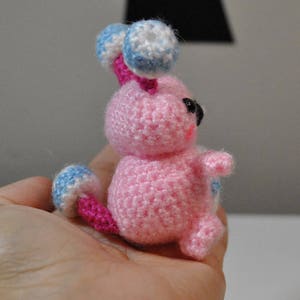 May include: A pink crocheted amigurumi toy with blue and white accents. The toy has a round body, two small arms, and a tail. The toy has black eyes and a pink nose.