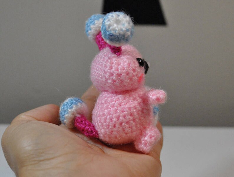 May include: A pink crocheted amigurumi toy with blue and white accents. The toy has a round body, two small arms, and a tail. The toy has black eyes and a pink nose.