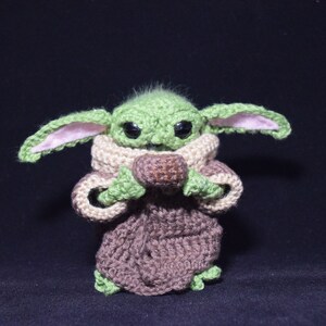 May include: A green crocheted plush toy with large eyes and brown ears. The toy is wearing a brown crocheted robe and holding a brown crocheted ball.
