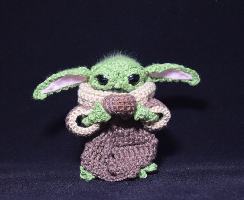 May include: A green crocheted plush toy with large eyes and brown ears. The toy is wearing a brown crocheted robe and holding a brown crocheted ball.