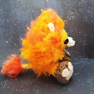 May include: A crocheted lion with orange and yellow fur, brown legs, and white paw pads. The lion is sitting on a dark background.