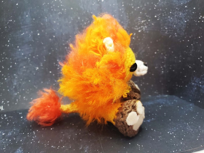 May include: A crocheted lion with orange and yellow fur, brown legs, and white paw pads. The lion is sitting on a dark background.