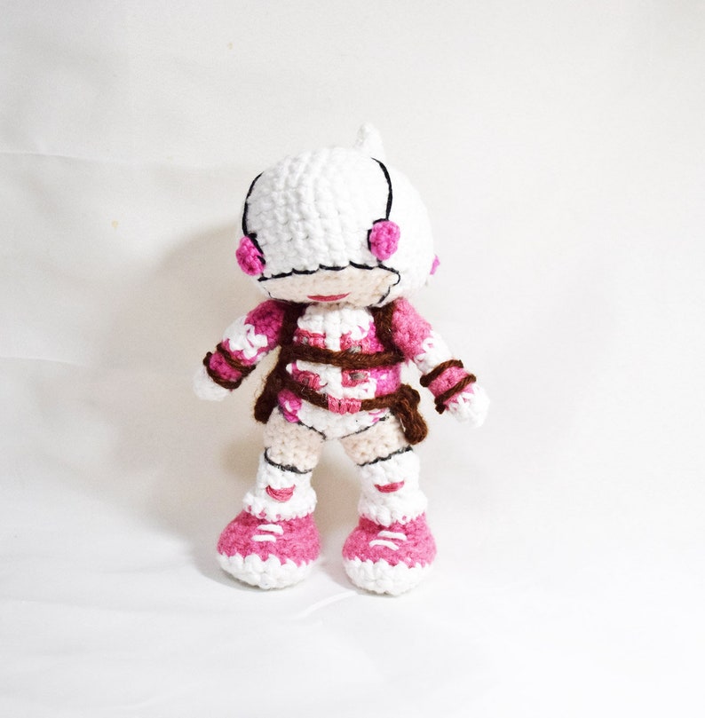 May include: A crocheted plush toy in the shape of a person wearing a white helmet with pink accents, a pink and white suit, and pink shoes.