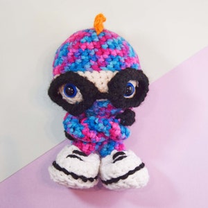 May include: A crocheted toy wearing a pink, blue and white patterned hat with a black eye mask and white trainers.