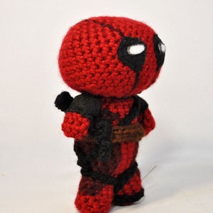 May include: A crocheted Deadpool character toy with a red and black costume. The toy has a white eye mask with black eyes and a brown belt.