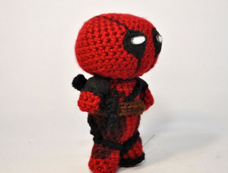 May include: A crocheted Deadpool character toy with a red and black costume. The toy has a white eye mask with black eyes and a brown belt.