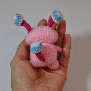 May include: A pink crocheted amigurumi toy with blue and white eyes and pink antennae. The toy is being held in a hand.