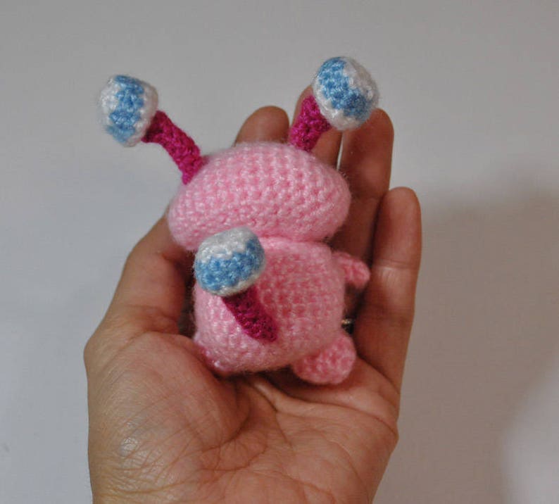 May include: A pink crocheted amigurumi toy with blue and white eyes and pink antennae. The toy is being held in a hand.