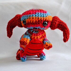 May include: A crocheted stuffed toy with red, orange, blue, and pink stripes. The toy has large, black eyes and a red, crocheted tail.