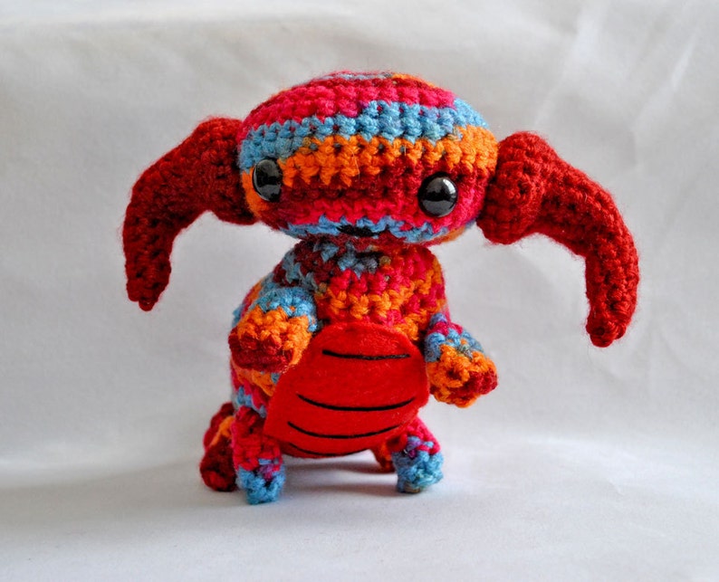 May include: A crocheted stuffed toy with red, orange, blue, and pink stripes. The toy has large, black eyes and a red, crocheted tail.
