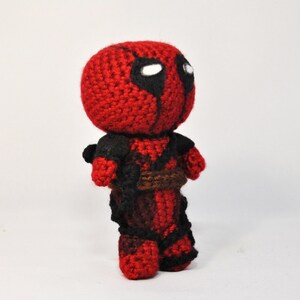 May include: A crocheted Deadpool plush toy with a red and black costume. The toy has a white eye mask and is standing on a white surface.