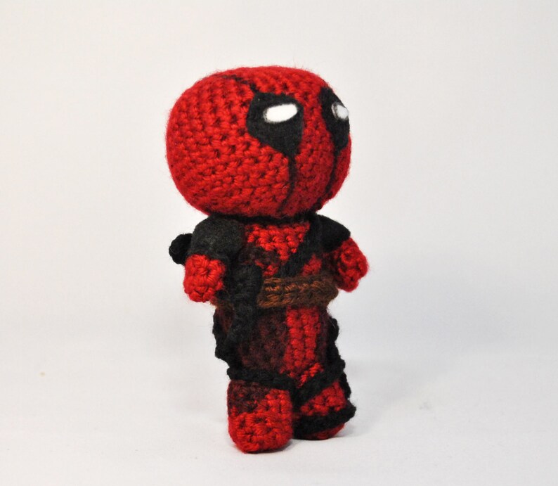 May include: A crocheted Deadpool plush toy with a red and black costume. The toy has a white eye mask and is standing on a white surface.