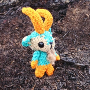 May include: A crocheted toy animal with blue and orange fur, resembling a goat or ram. The toy has a long, curled orange horn and a white face with black eyes.