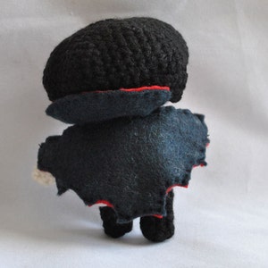 May include: A crocheted plush toy with a black felt cape and red trim. The toy has a black crocheted head and a black felt body.