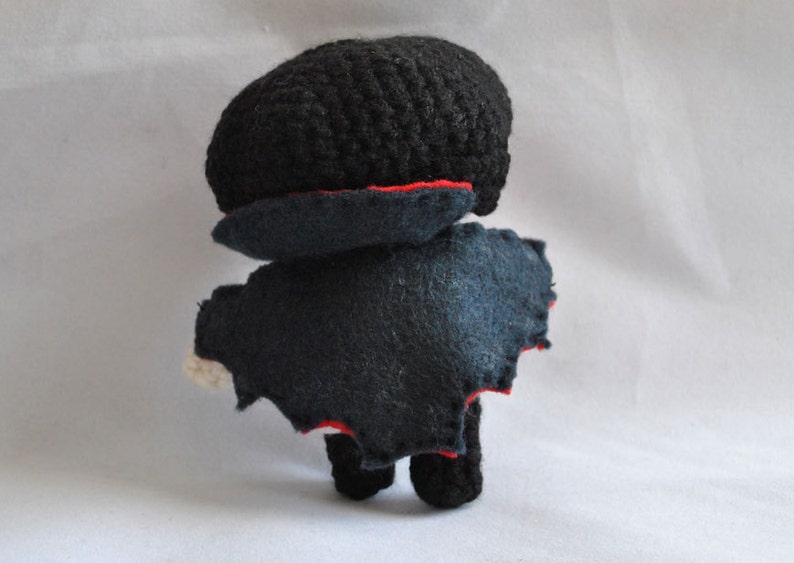 May include: A crocheted plush toy with a black felt cape and red trim. The toy has a black crocheted head and a black felt body.