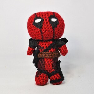 May include: A red and black crocheted Deadpool character with a white eye mask. The character is standing on a white surface.