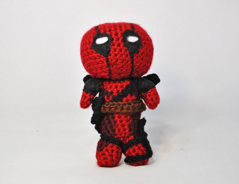 May include: A red and black crocheted Deadpool character with a white eye mask. The character is standing on a white surface.