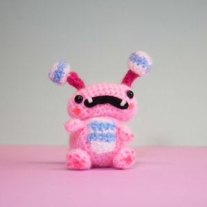 May include: A pink crocheted toy with a black moustache, blue and white striped body, and two blue and white striped antennae. The toy is sitting on a pink surface.