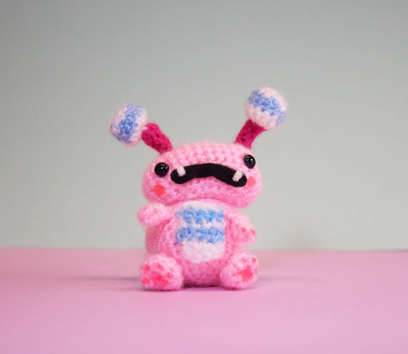 May include: A pink crocheted toy with a black moustache, blue and white striped body, and two blue and white striped antennae. The toy is sitting on a pink surface.