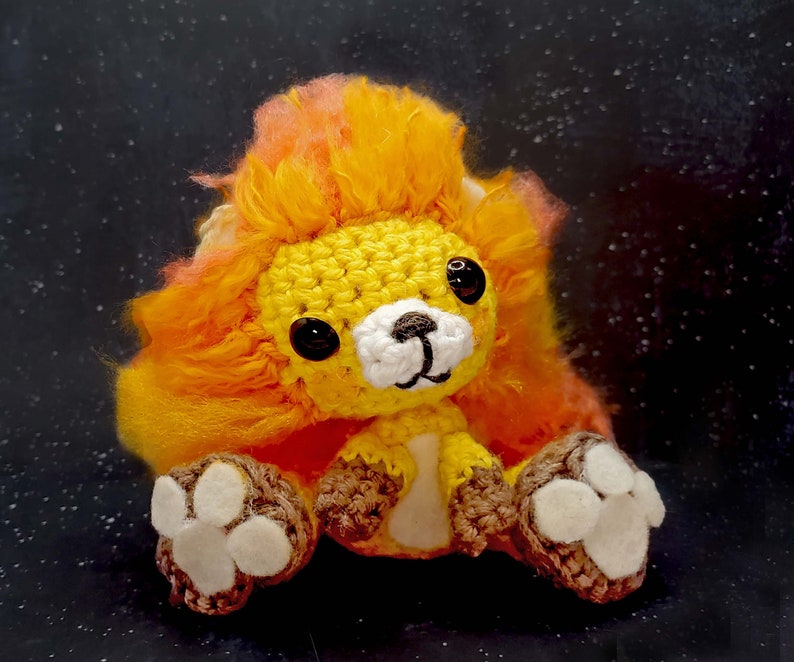 May include: A crocheted lion plush toy with a yellow and orange mane. The toy has white paws and a white nose.
