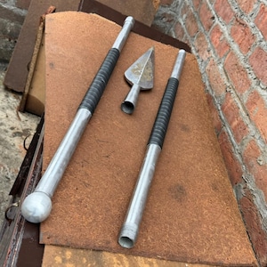 May include: Three metal spear parts are displayed on a brown surface. Two long, silver shafts have black grips. A silver arrowhead is also present. The spear parts are arranged against a brick wall.