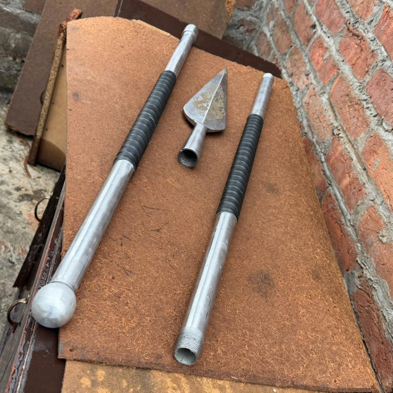 May include: Three metal spear parts are displayed on a brown surface. Two long, silver shafts have black grips. A silver arrowhead is also present. The spear parts are arranged against a brick wall.