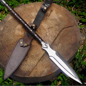 Op de afbeelding: Een handgemaakte speer met een donkere houten schacht en een zilveren blad. De speer wordt gecombineerd met een bruine leren schede en een zwarte leren riem. De speer ligt op een houten oppervlak.