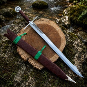 Op de afbeelding: Een stalen zwaard met een zilverkleurige pareerstang en een bruin, met leer omwikkeld handvat met een ronde pommel. Het lemmet heeft een geëtst patroon bij het handvat. Een bijpassende bruine lederen schede met groene koordaccenten en een zilveren punt.