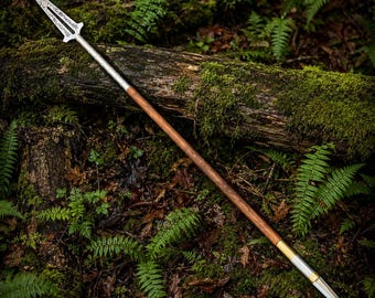 May include: A long spear featuring a sharp, double-edged metal blade with intricate etched patterns at the top. The shaft is made of smooth brown wood, connected to the blade and a pointed metal base by polished silver and gold-toned metal rings.