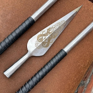 May include: A close-up of three spearheads. The central spearhead is silver with a gold-colored engraved design. The handles are silver with black leather grips. The spearheads are on a brown surface.