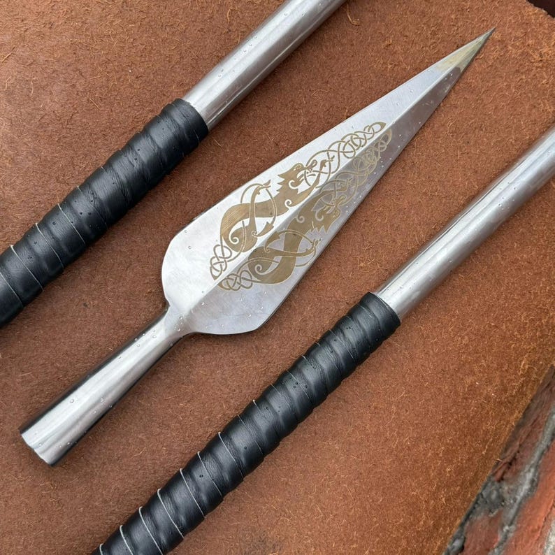 May include: A close-up of three spearheads. The central spearhead is silver with a gold-colored engraved design. The handles are silver with black leather grips. The spearheads are on a brown surface.