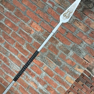 May include: A silver spear with a decorative blade featuring an engraved design. The handle is wrapped in black material for grip. The spear is set against a red brick wall.