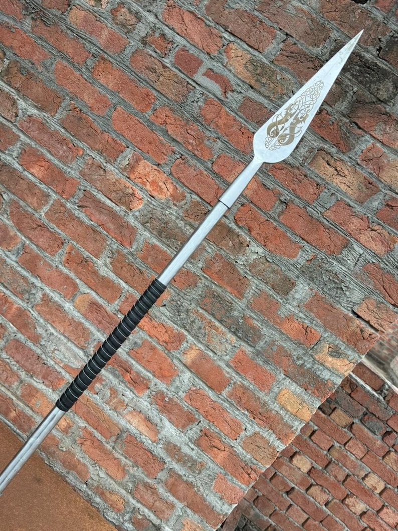 May include: A silver spear with a decorative blade featuring an engraved design. The handle is wrapped in black material for grip. The spear is set against a red brick wall.