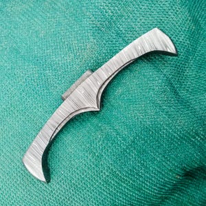 May include: A curved, Damascus steel knife blade with a textured pattern. The blade has a pointed tip and a decorative, notched spine. The knife is set against a green background.