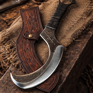 May include: A curved knife with a silver blade and a black handle, accompanied by a brown leather sheath. The knife features a ring at the handle's end. The sheath is adorned with carvings. The items are displayed on a wooden surface.