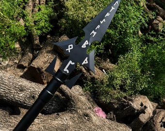 May include: A black spear with a sharp, pointed head featuring cut-out sections and white runic symbols engraved along the blade. The spear has a long, smooth, cylindrical black shaft that extends downwards from the decorative metal head.